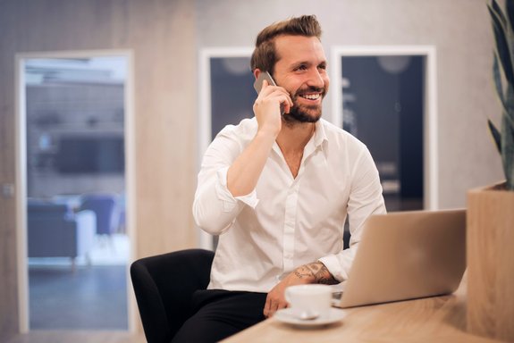 Ein lächelnder Mann in einem weißen Hemd sitzt an einem Tisch, hält ein Smartphone am Ohr und arbeitet an einem Laptop. Neben ihm steht eine Tasse Kaffee