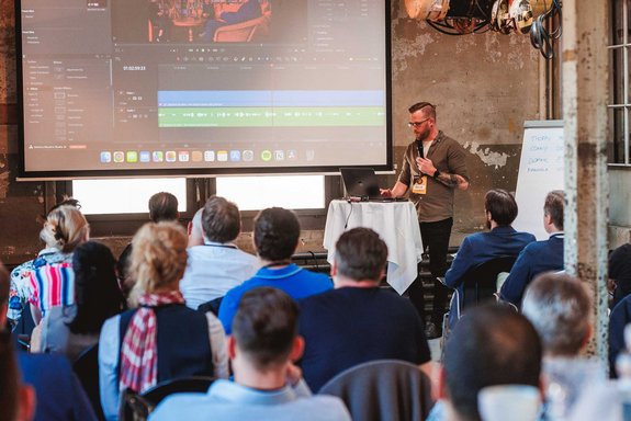 Felix Schmidt von Felix Content Marketing präsentiert eine Keynote in Zürich über KI in der Videobearbeitung. Er steht vor einer Leinwand, spricht ins Mikrofon und hat einen Laptop auf einem Tisch neben sich